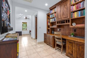Uma sala de estudos com escrivaninha de madeira, estantes, uma cadeira e janela com vista.