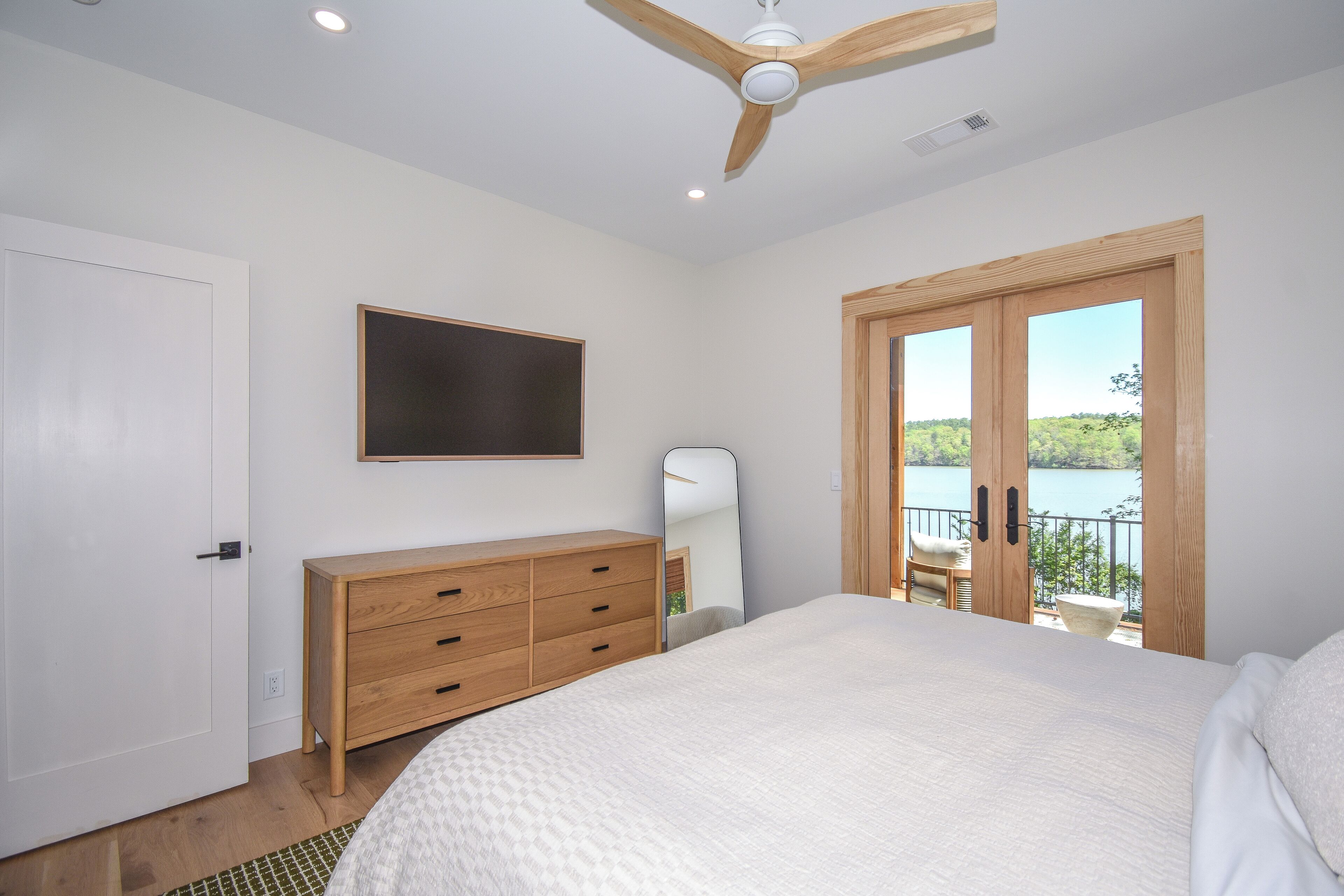 A bedroom with a large bed, wooden dresser, ceiling fan, and a view of a lake through a sliding door.