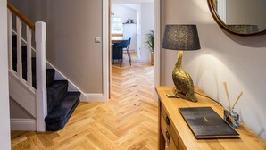 Un pasillo con suelo de madera, una escalera con barandillas blancas, un escritorio con lámpara y un espejo en la pared.