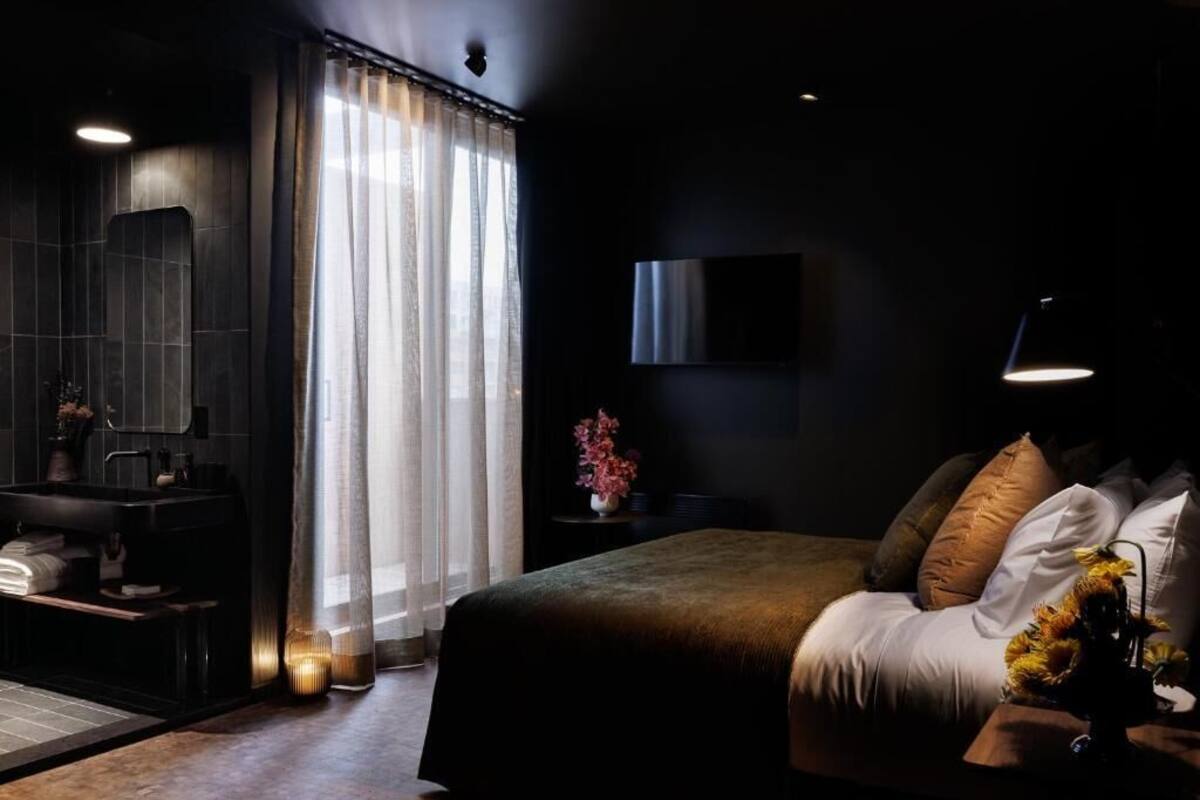 A modern hotel room with a dark color scheme, a large bed, a bedside table with a lamp, and a view of the city through sheer curtains.