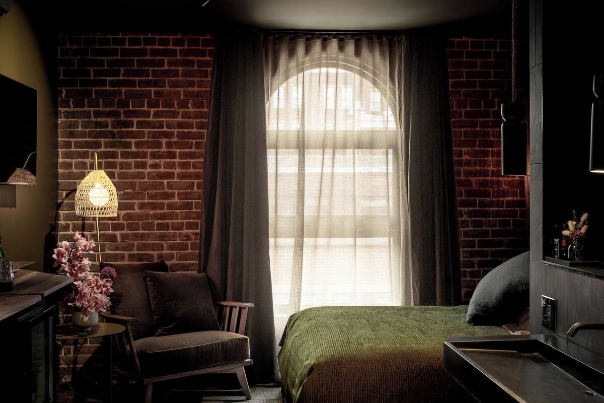A dimly lit room with a brick wall, a bed with a green blanket, a small table with a lamp, and a chair.