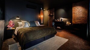 A modern bedroom with a large bed, a side table with a lamp, a brick accent wall, and a small shelf with decorative items. - Maison Grinder (Montréal)