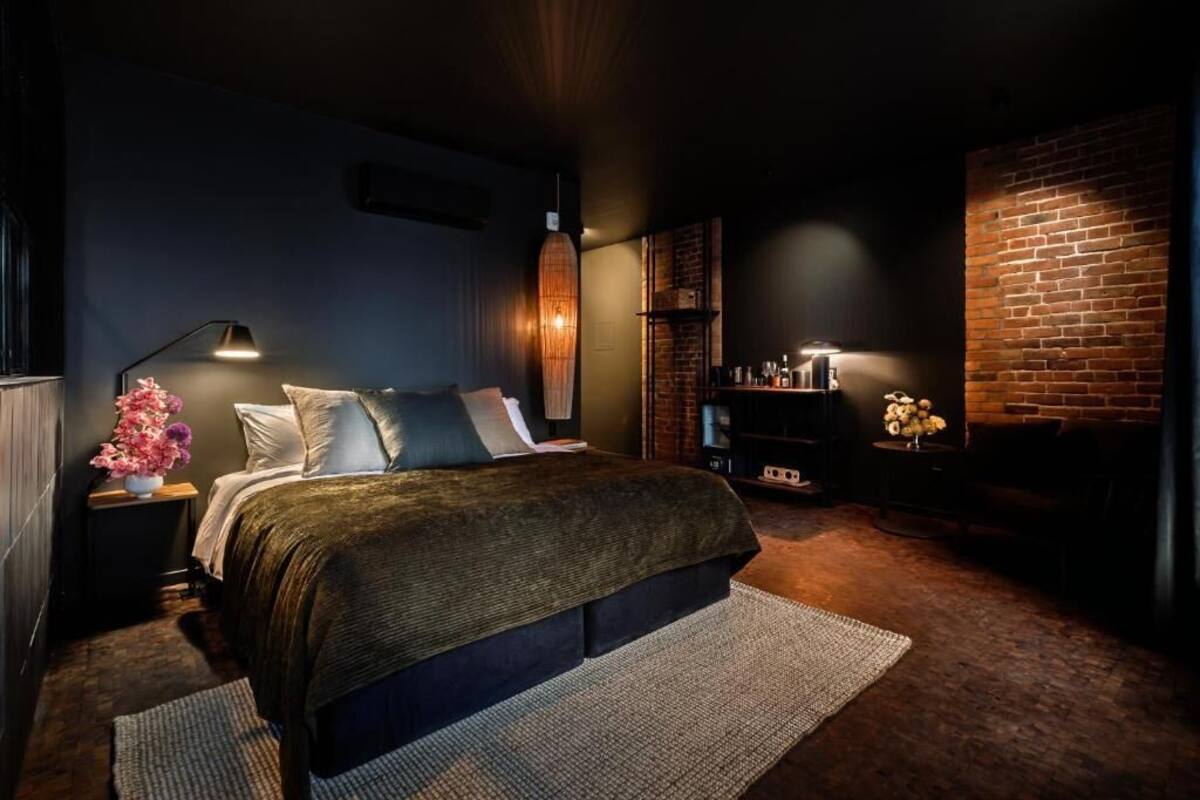 A modern bedroom with a large bed, a side table with a lamp, a brick accent wall, and a small shelf with decorative items.