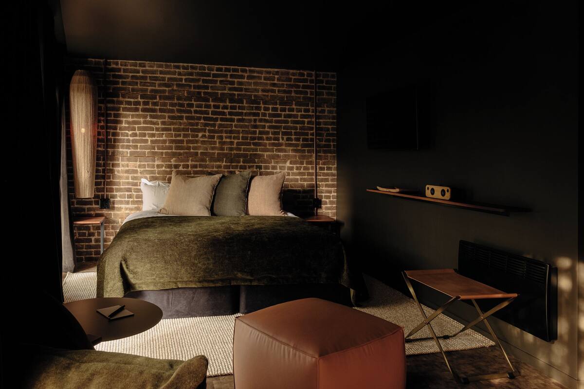 A bedroom with a brick wall, a bed with pillows, a bedside table, a small wooden shelf, a folding chair, and a radiator.