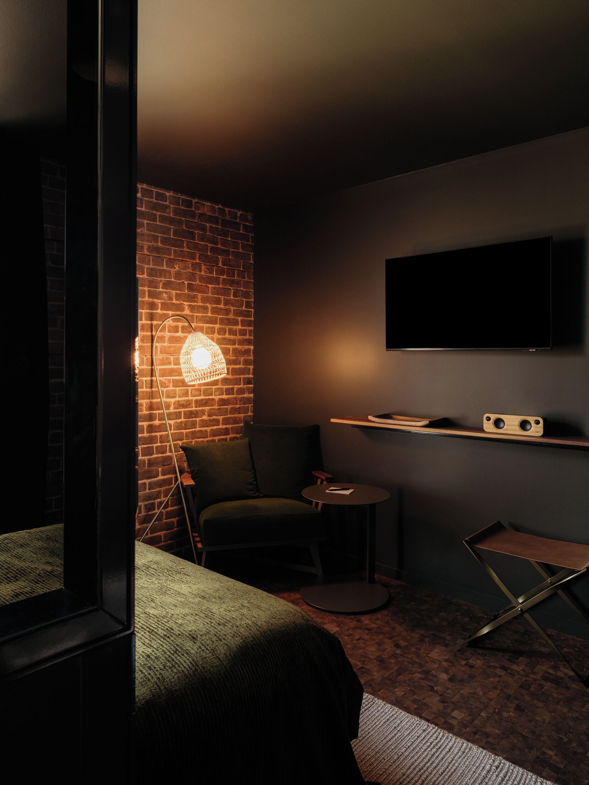 Una habitación con poca luz, pared de ladrillo, televisor de pantalla plana montado en la pared, un sillón verde, una mesita redonda y una silla plegable.