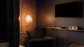 Una habitación con poca luz, pared de ladrillo, televisor de pantalla plana montado en la pared, un sillón verde, una mesita redonda y una silla plegable.