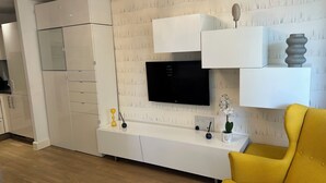A modern living room with a flat-screen TV mounted on a textured wall, a white entertainment unit with a vase and a plant, and a yellow armchair. - 1-Bed Apt, Sleeps 4, Near Attractions & Transport (Lewisham)