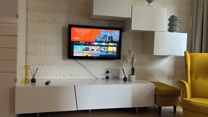 A modern living room with a flat-screen TV mounted on the wall, a white entertainment unit, a yellow armchair, and a small table with decorative items. - 1-Bed Apt, Sleeps 4, Near Attractions & Transport (Lewisham)