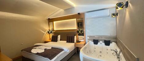 A hotel room with a bed, a bedside table, a nightstand, a large mirror, a wall-mounted light fixture, and a bathtub.