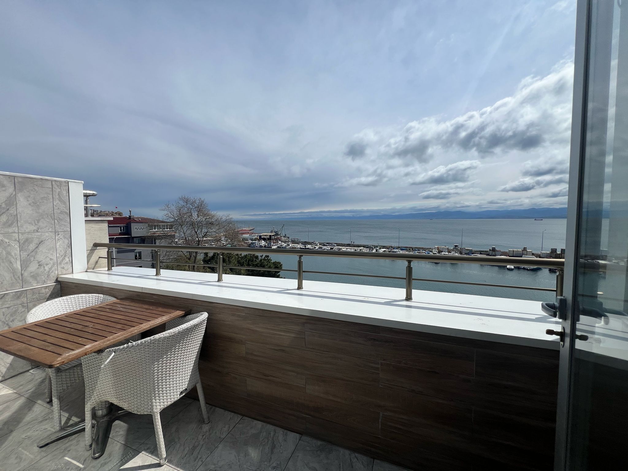 A balcony with a wooden table and wicker chair, overlooking a marina and distant mountains.