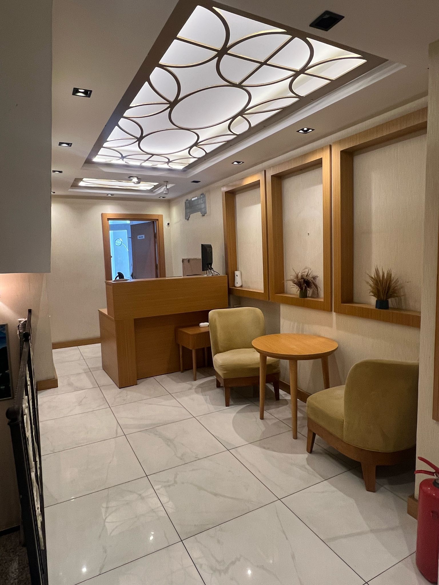 A hotel lobby with a reception desk, two armchairs, a small table, and a ceiling light fixture.