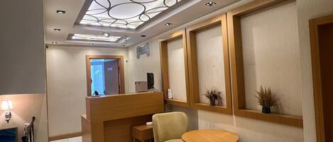A hotel reception area with a wooden desk, two chairs, a small table, and a decorative ceiling light fixture.