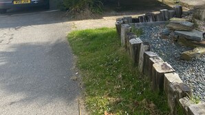 Une maison mobile avec un garage, une voiture garée devant, et une zone herbeuse délimitée par une clôture en bois et des rochers.