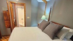 Un dormitorio con estructura de cama de madera, colchón blanco y dos almohadas. Hay una puerta de madera y una lámpara sobre una mesita de noche.