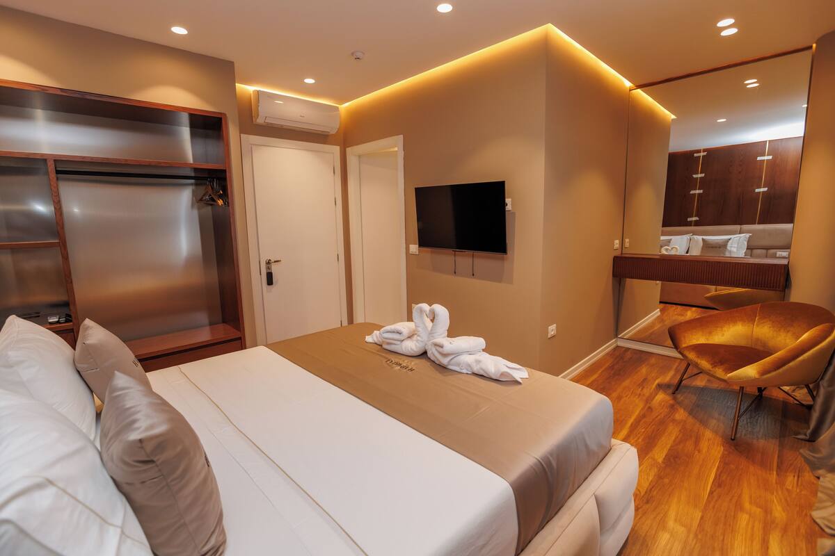 A modern hotel room with a large bed, a TV mounted on the wall, a wooden floor, and a seating area with a chair.