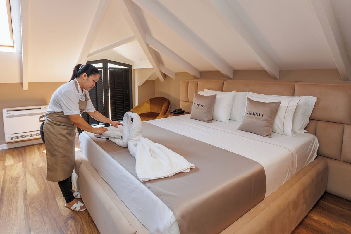 A hotel room with a large bed, a wooden floor, a ceiling with exposed beams, and a person folding a towel.