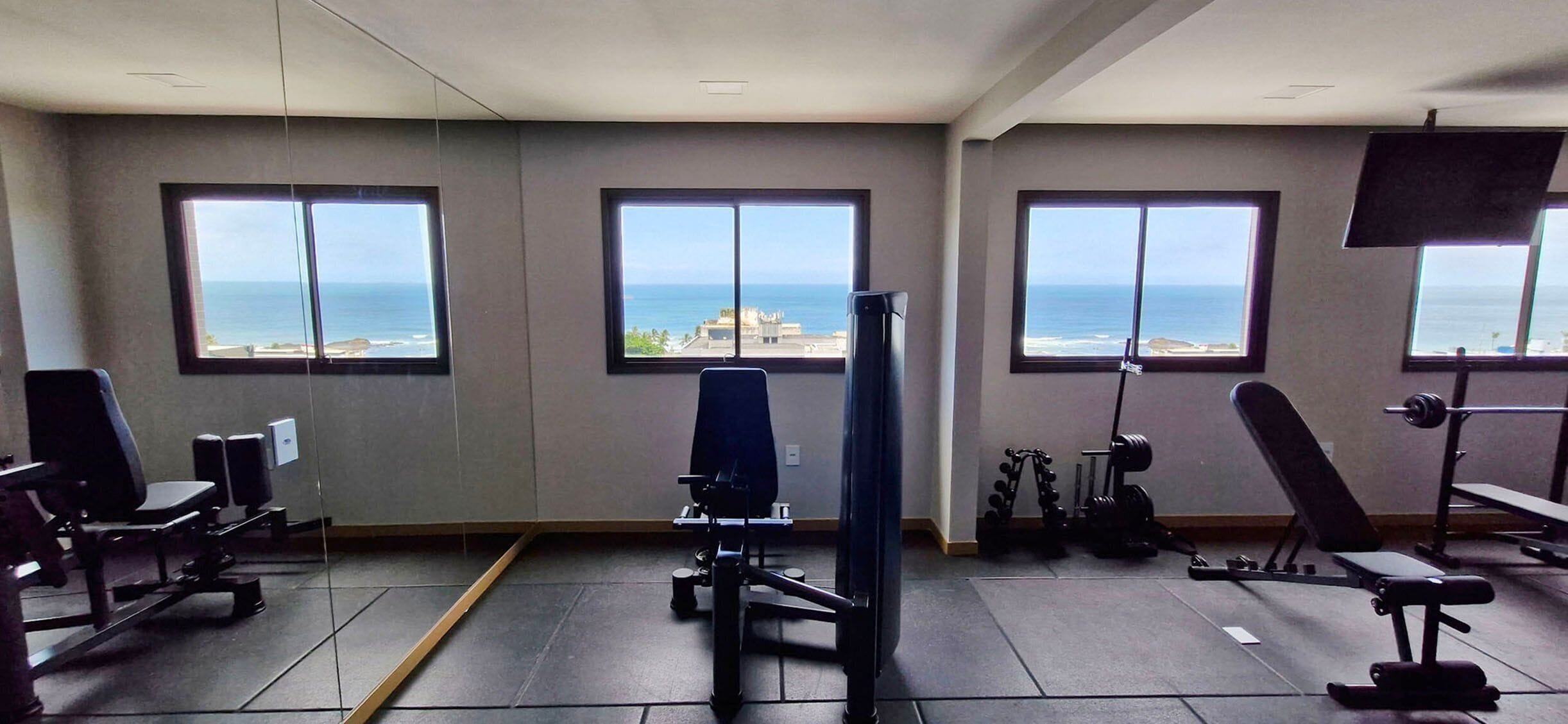 Academia com amplas janelas de frente para o oceano, equipada com diversos aparelhos de exercícios.