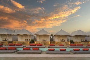 A row of white tents with lounge chairs and red patterned cushions, set against a sunset sky.