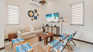 A living room with a sofa, coffee table, and two armchairs. There is a TV mounted on the wall, a ceiling fan, and a window with blinds. - Stylish Condo in the Heart of Las Catalinas with Vibrant Design & Exclusive Beach Club Access (Las Catalinas)
