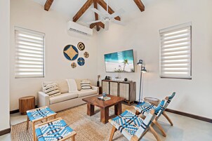 A living room with a sofa, coffee table, and two armchairs. There is a TV mounted on the wall, a ceiling fan, and a window with blinds.