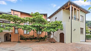 A courtyard with a brick floor, a tree with green leaves, and a building with a light-colored facade and a wooden balcony. - Stylish Condo in the Heart of Las Catalinas with Vibrant Design & Exclusive Beach Club Access (Las Catalinas)