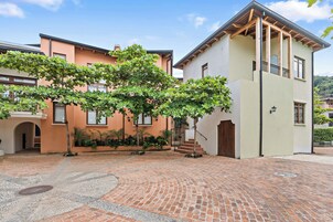 Un patio con suelo de ladrillo, un árbol de hojas verdes y un edificio con fachada clara y un balcón de madera.