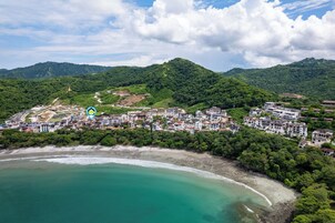 Vista aérea de un pueblo costero con playa, vegetación densa y una combinación de edificios residenciales.