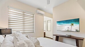 A hotel room with a bed, a TV on a wooden stand, and a large window with blinds. - Stylish Condo in the Heart of Las Catalinas with Vibrant Design & Exclusive Beach Club Access (Las Catalinas)