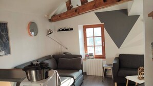 Tunnelmallinen olohuone, jossa on moderni keittiöalue, kattopalkit, suuri ikkuna ja takka.