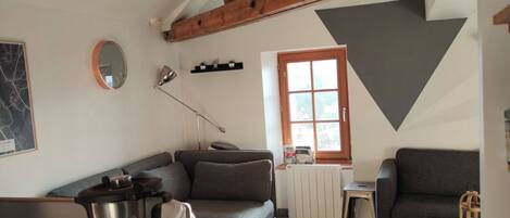 A cozy living room with a modern kitchen area, wooden beams on the ceiling, a large window, and a fireplace.