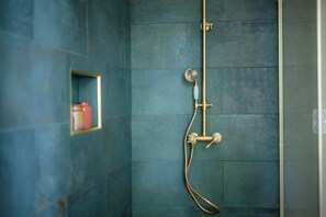 A bathroom with a blue tiled wall, a showerhead, and a small shelf with toiletries. - Château de Sienne (Chusclan)