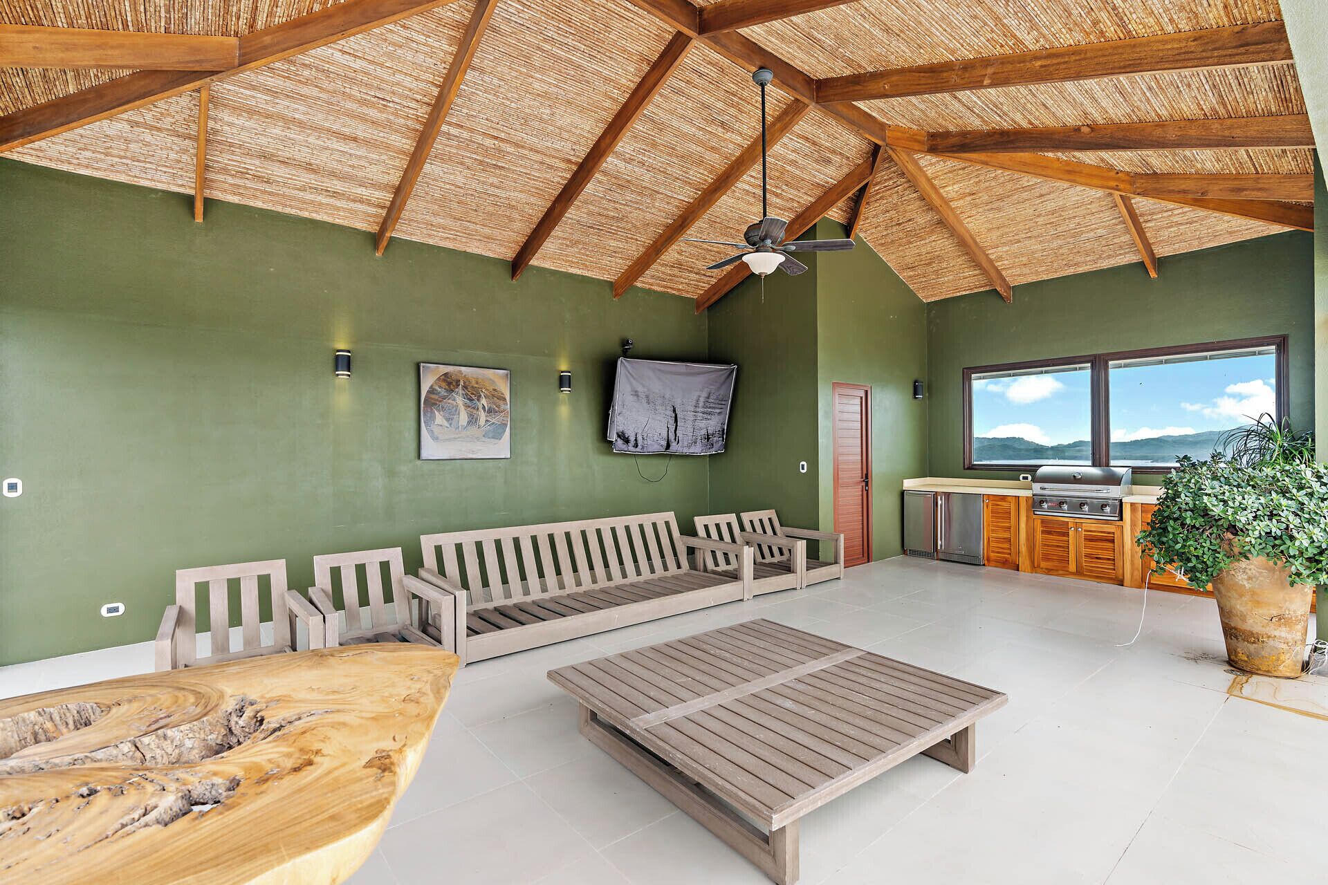 Ein geräumiges Zimmer mit hoher Decke, einem Holzboden, einem Deckenventilator, einer hölzernen Sitzbank, einem Holztisch, einer Küchenzeile mit Grill, einem Fenster mit Blick und zwei Wandleuchten.
