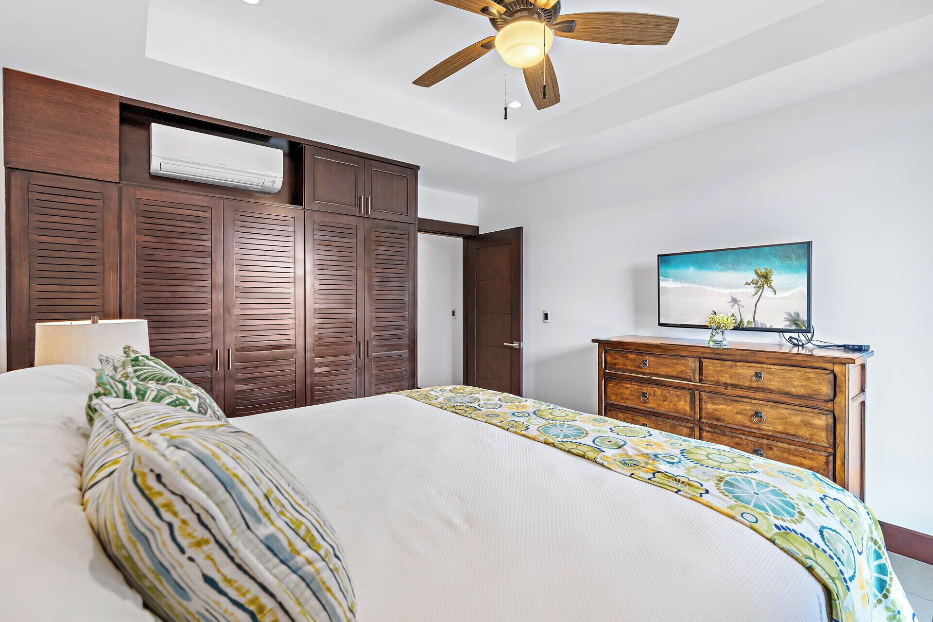 A bedroom with a bed, a wooden wardrobe, a television on a wooden dresser, and a ceiling fan.
