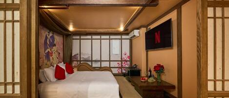 A hotel room with a bed, a TV mounted on the wall, a small table with a vase of flowers, and a window with a view of mountains.