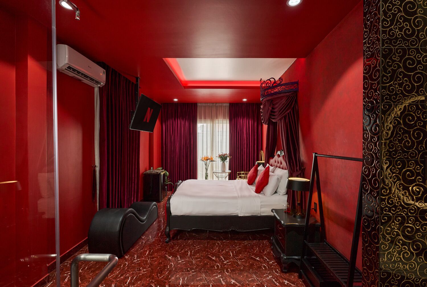 A hotel room with a red theme, including red walls, a red carpet, and red curtains. There is a bed with white bedding and red pillows, a black armchair, a small table with a lamp, and a TV mounted on the wall.