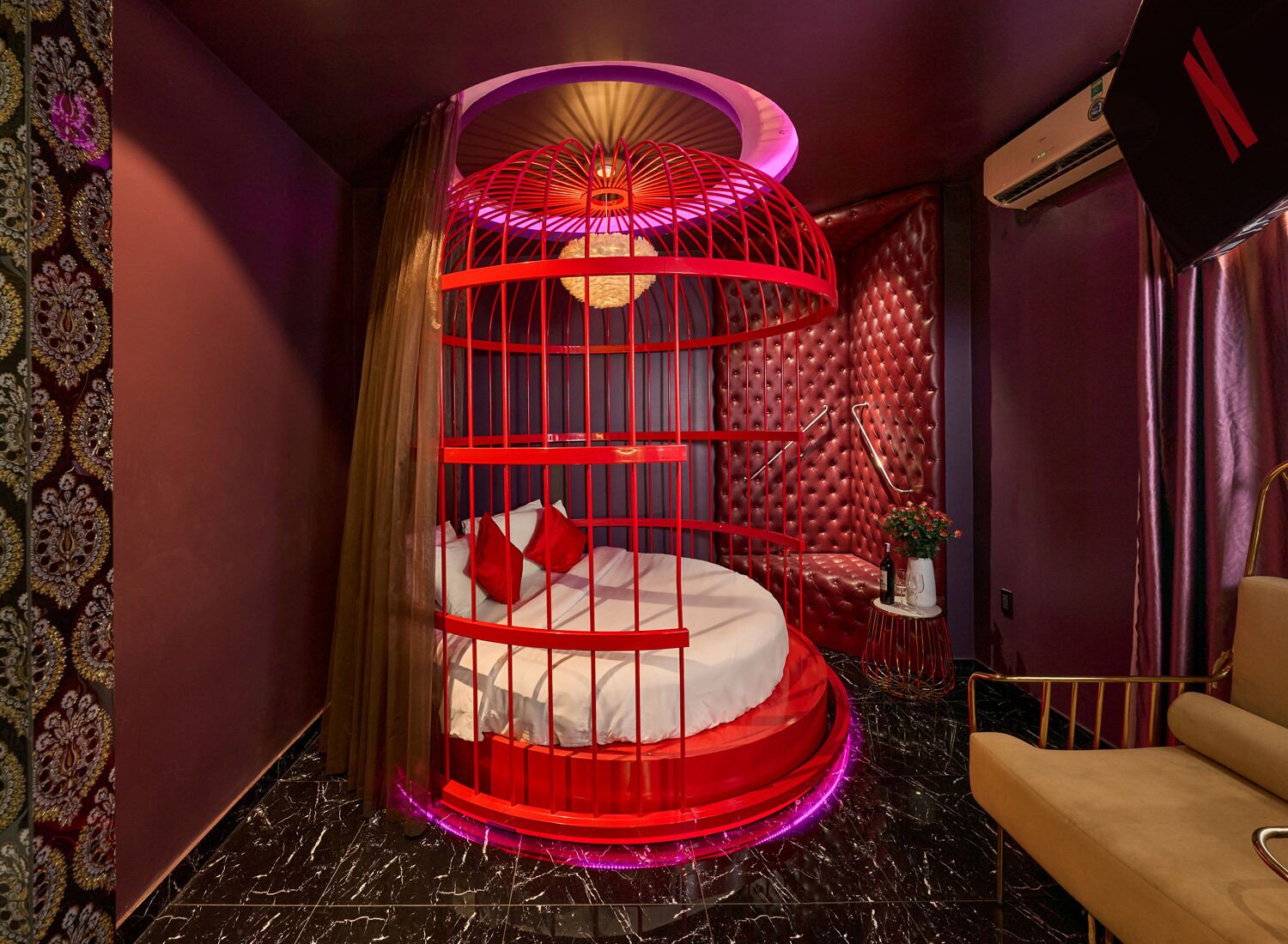 A circular bed with a red frame and a canopy, a tufted leather headboard, a small side table with a vase, and a wall-mounted air conditioner.