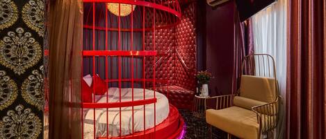 Une chambre à coucher luxueuse avec un lit rond dont le cadre est rouge, une tête de lit rouge à motifs capitonnés, un fauteuil à armatures dorées et un téléviseur moderne.