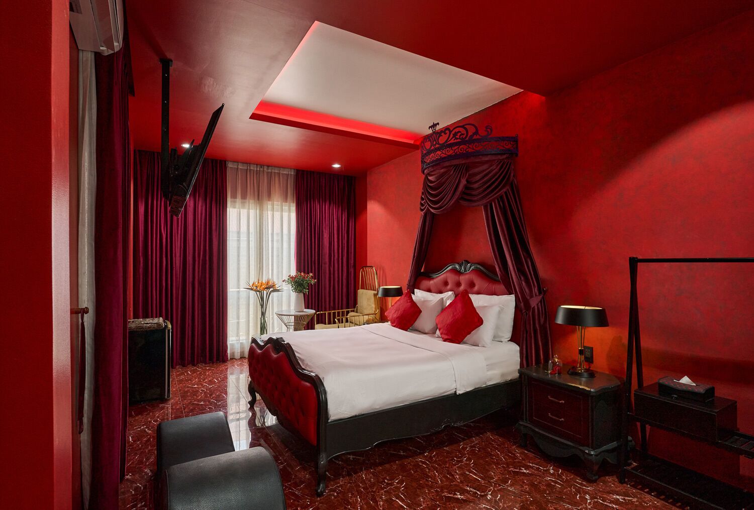 A red-themed bedroom with a large bed, a canopy, and a nightstand with a lamp.