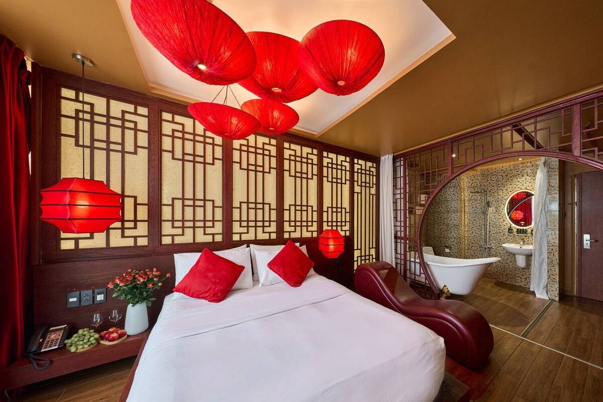 A hotel room with a large bed, red pillows, a red hanging lamp, a wooden partition with a geometric design, and a round mirror with a red frame.