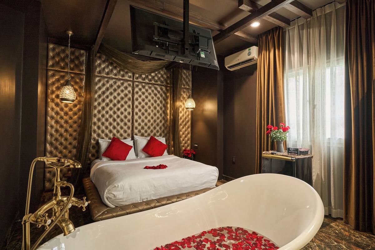A luxurious hotel room with a large bed, a separate smaller bed, a bathtub filled with rose petals, a gold bathtub faucet, and a window with curtains.