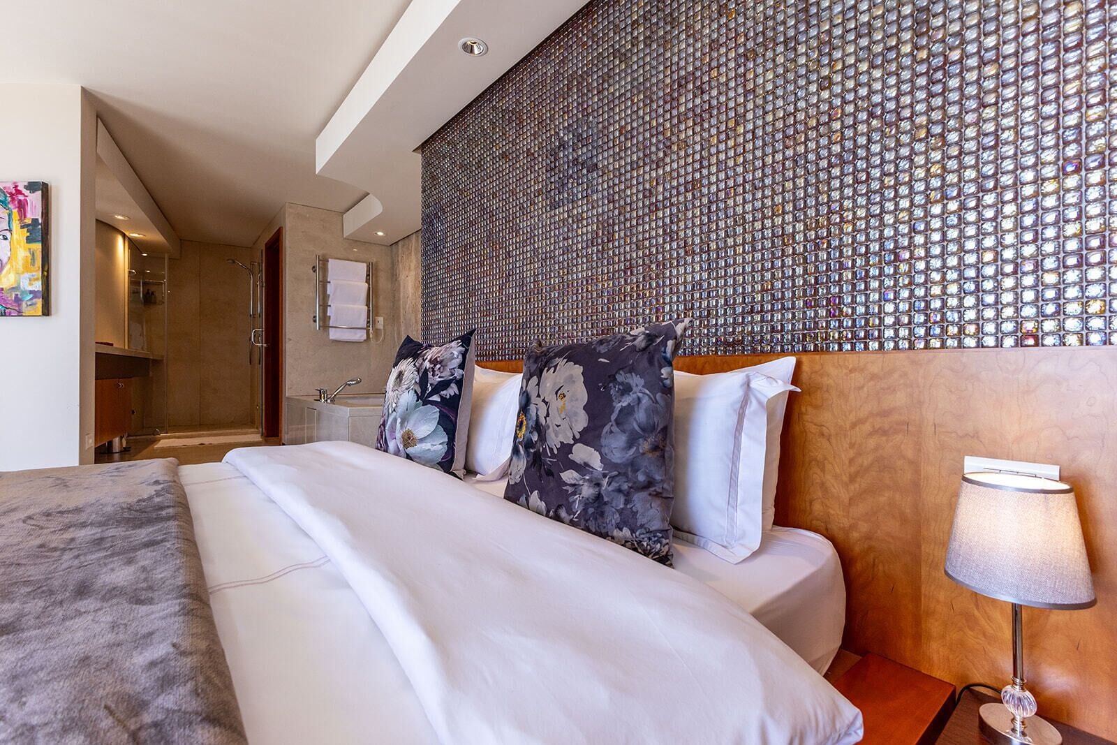 A hotel room with a large bed, a wooden headboard, a decorative wall, a bathroom with a shower, and a painting on the wall.