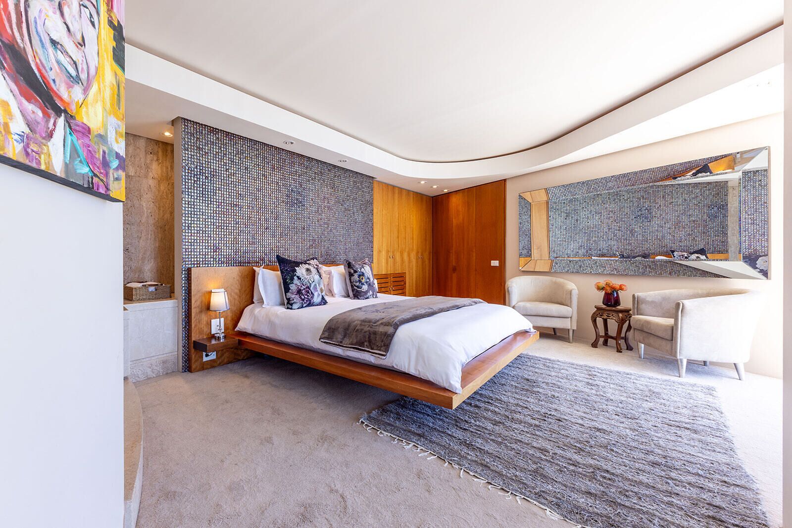 A modern bedroom with a large bed, a wooden headboard, a seating area with a chair and a small table, and artwork on the walls.