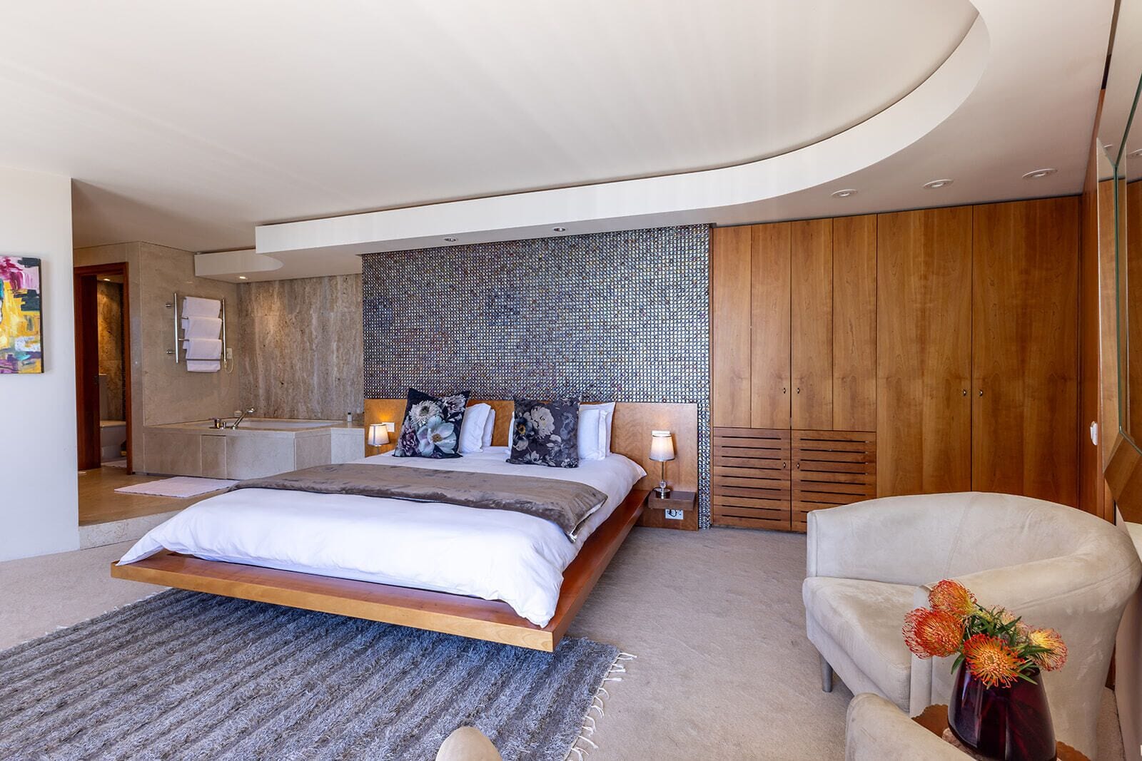 A modern bedroom with a large bed, wooden wardrobe, and a bathroom with a mirror and towel rack.