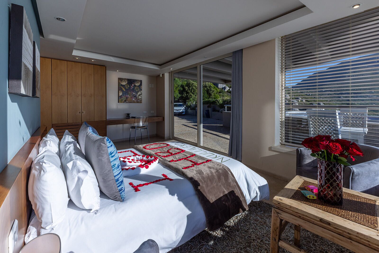 A modern bedroom with a large bed, a desk, and a view of the outdoors through large windows.