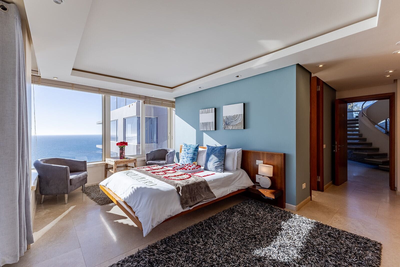 A modern bedroom with a large bed, a view of the ocean, a sitting area with a chair and table, and a staircase leading to another level.