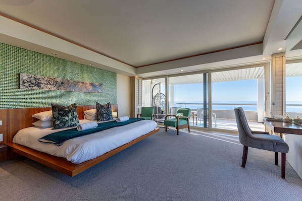 A spacious hotel room with a large bed, a balcony with a view of the ocean, and a seating area with chairs and a table. - The Marvel Beach House (Cape Town)