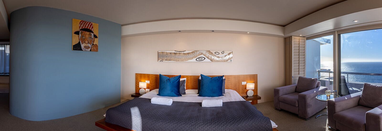 Une chambre d’hôtel moderne avec un plafond incurvé, un grand lit avec une literie bleue et blanche, deux lampes de chevet et une vue sur l’océan par de grandes fenêtres.