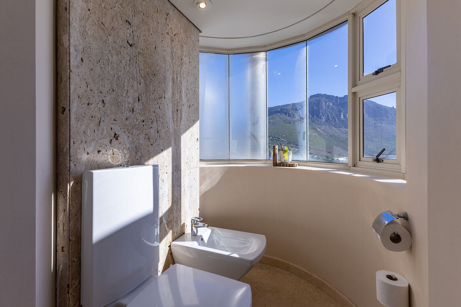 Une salle de bain équipée d’une baignoire, de toilettes et d’un lavabo, de grandes fenêtres offrant une vue sur les montagnes et d’un porte-rouleau de papier toilette.