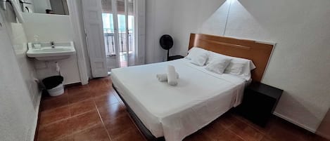 Ein Hotelzimmer mit einem Bett, einem Nachttisch, einer Wandleuchte, einem Fenster mit Jalousien sowie einem Badezimmer mit Waschbecken und Spiegel.