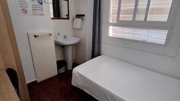 A small room with a bed, a sink, a mirror, a television, and a window with blinds.